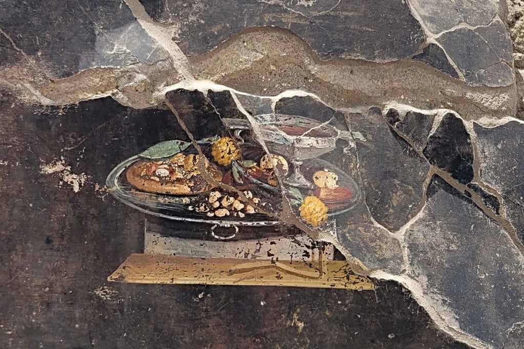 El fresco representa una bandeja de plata con variedades de frutas, especias, una copa de vino rojo y una masa redonda, similar a una pizza.