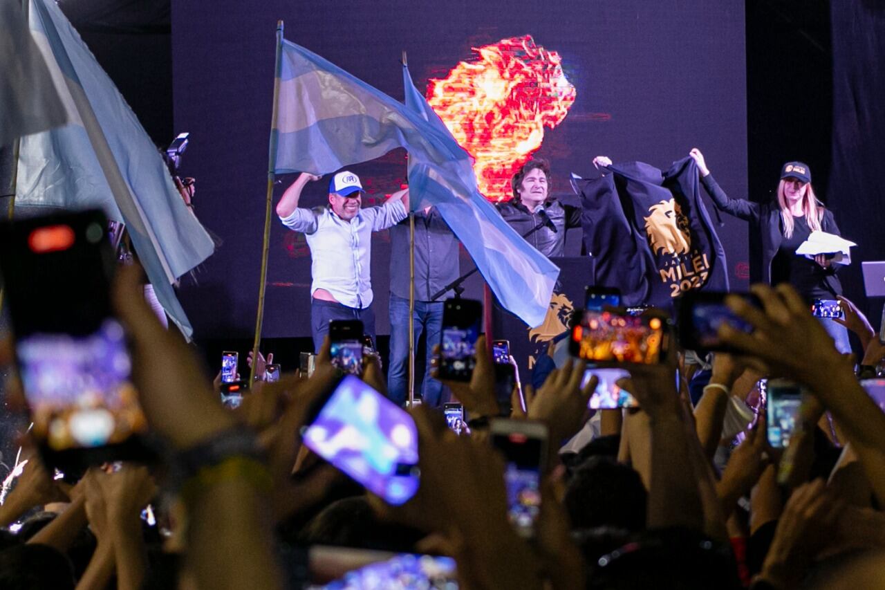 Frente a las banderas argentina, Ricardo Bussi y Javier Milei saludan en el escenario con los brazos en alto.