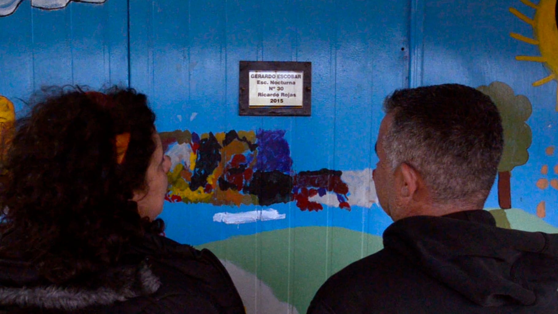 Ana Lucas y Adrián Gómez, docentes, frente a la placa que recuerda a Pichón en su escuela.