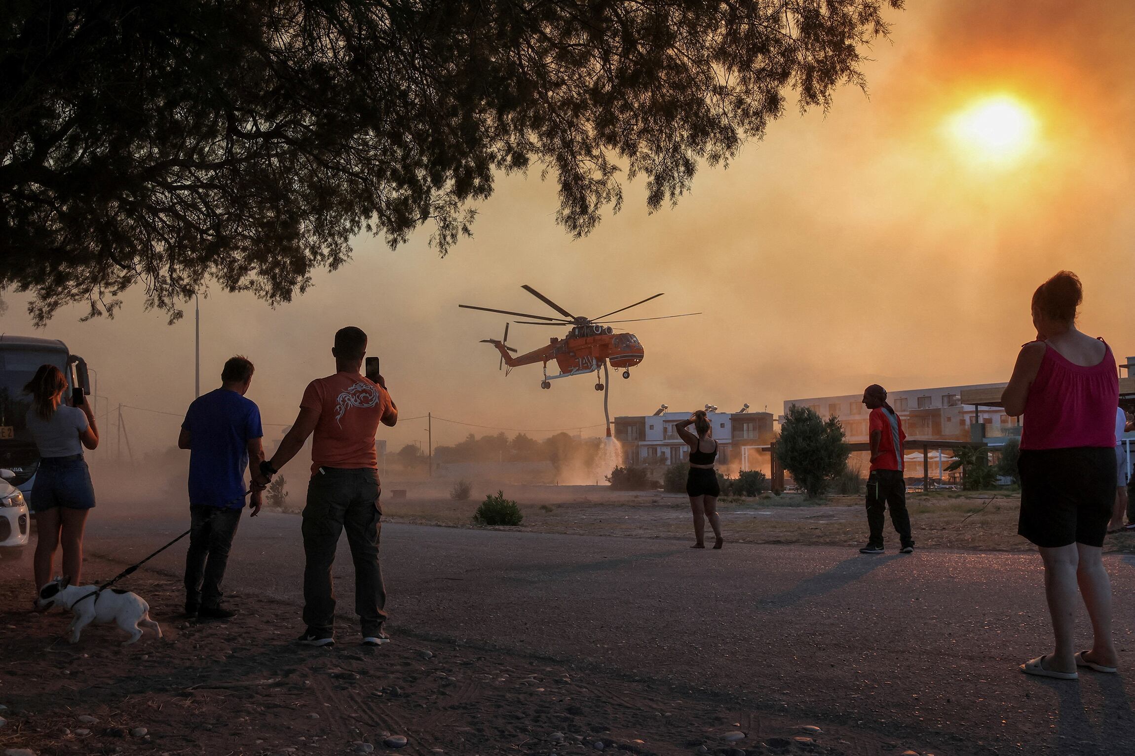 Varias personas observan un helicóptero de bomberos llenando de agua un estanque, mientras arde un incendio en el pueblo de Gennadi, en la isla de Rodas, Grecia. 