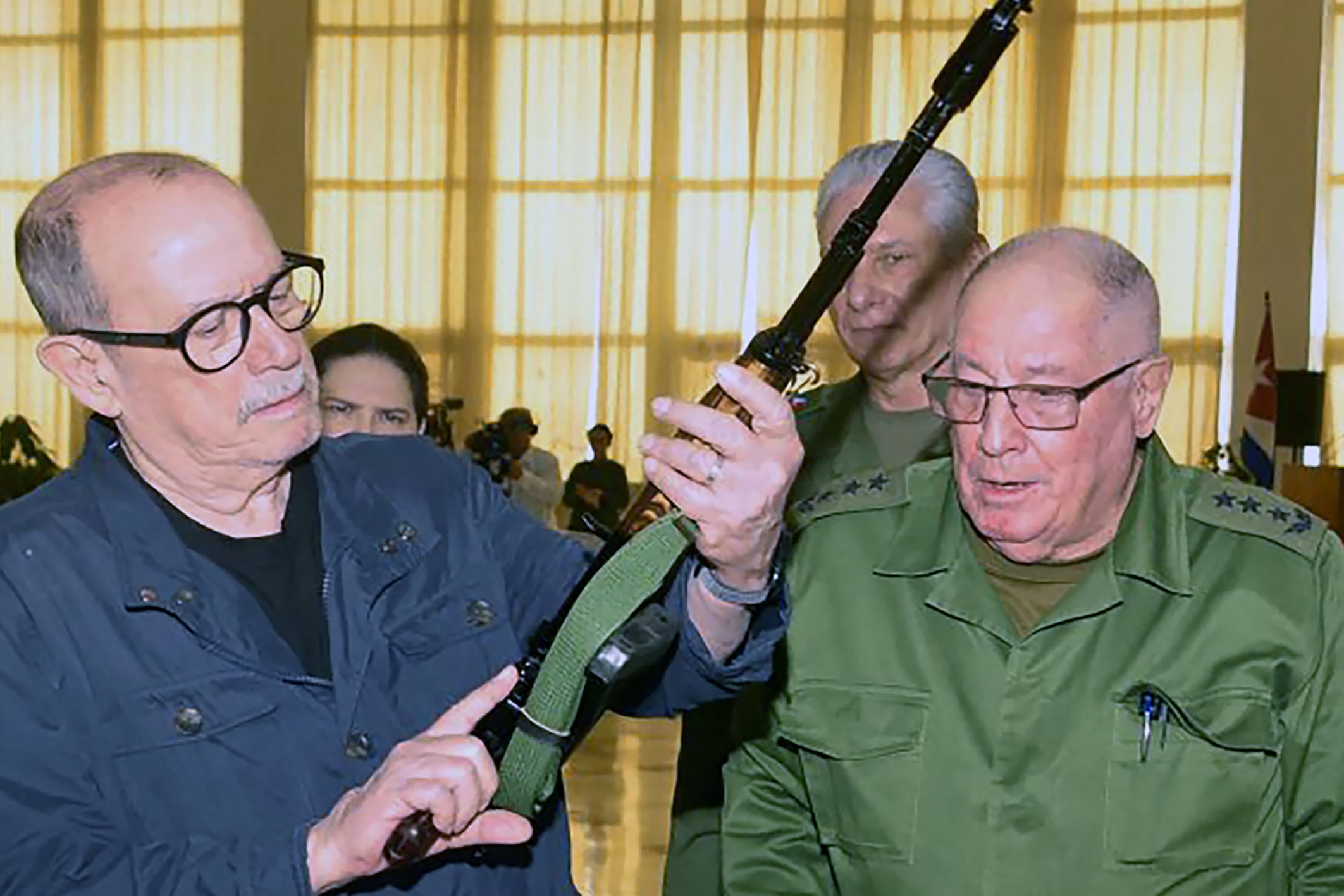 Handout picture released by Cuban website Cubadebate showing Cuban singer-songwriter Silvio Rodriguez (L) holding an AKM rifle handed to him by the Minister of the Revolutionary Armed Forces of Cuba, Army Corps General Alvaro Lopez Miera and President Miguel Diaz-Canel (background), during a ceremony in the framework of the country's National Defence Day, in Havana on March 20, 2026, days after he expressed his will in case the United States were to carry out its threat to invade the island nation. Rodriguez, 79, received the weapon and a replica in response to the request he made on March 18, when he stated on his blog Segunda Cita: "I demand my AKM, if they come at us. And let it be clear that I mean this very seriously." (Photo by Handout / CUBADEBATE / AFP) / RESTRICTED TO EDITORIAL USE - MANDATORY CREDIT "AFP PHOTO / CUBADEBATE / HANDOUT " - NO MARKETING NO ADVERTISING CAMPAIGNS - DISTRIBUTED AS A SERVICE TO CLIENTS