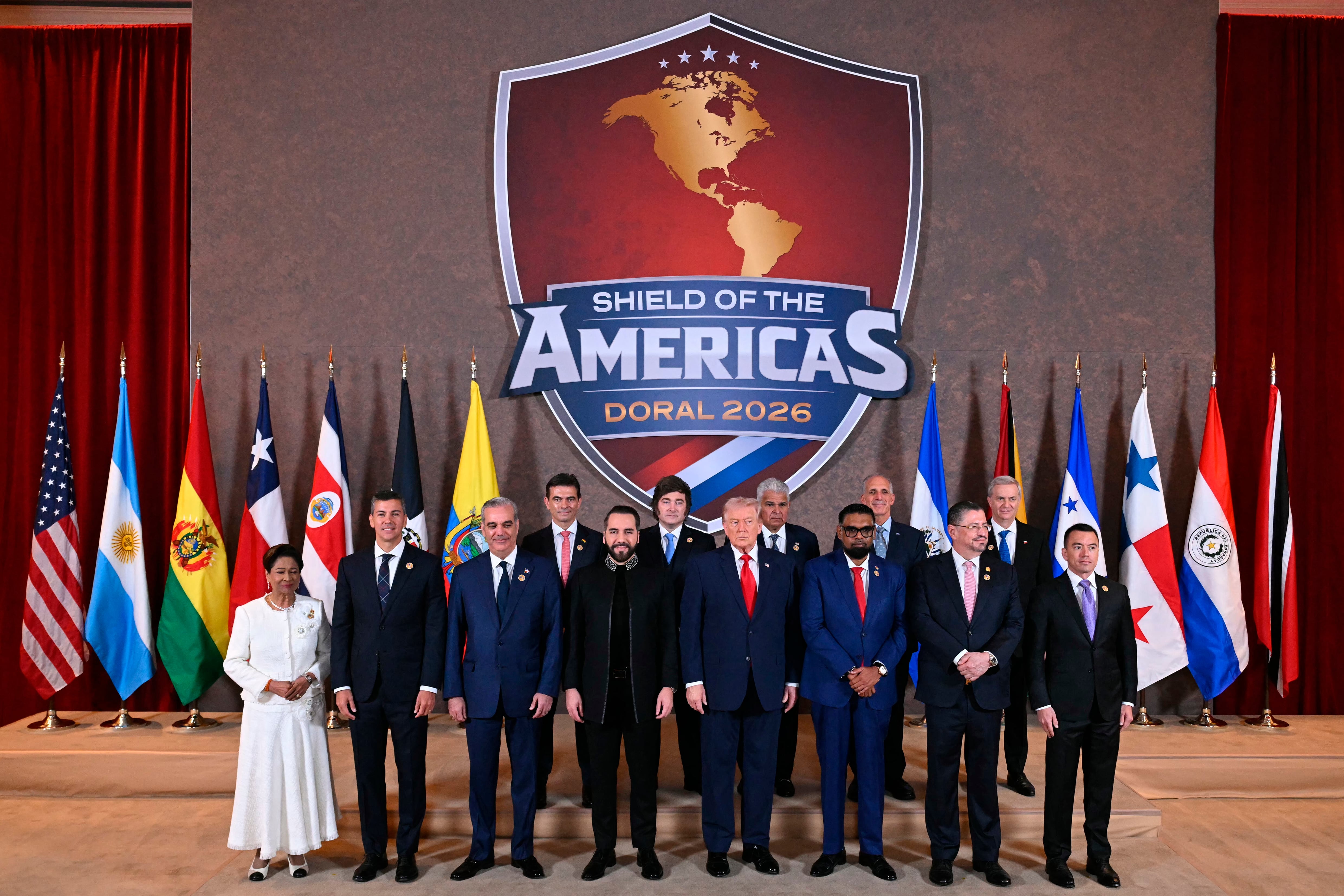 US President Donald Trump (C) stands with, (L/R), Trinidad and Tobago's Prime Minister Kamla Persad-Bissessar, Santiago Peña, President of the Republic of Paraguay, Luis Abinader, President of the Dominican Republic, Rodrigo Paz Pereira, President of Bolivia, Nayib Bukele, President of El Salvador, Javier Milei, President of Argentina, José Raúl Mulino, President of Panama, Mohamed Irfaan Ali, President of Guyana, Nasry “Tito” Asfura, President of Honduras, Rodrigo Chaves Robles, President of Costa Rica, José Antonio Kast, President-elect of Chile, and Daniel Roy Gilchrist Noboa Azín, President of Ecuador, during a group photograph at the start of the "Shield of the Americas" Summit at Trump National Doral in Miami, Florida, March 7, 2026. President Trump is hosting a dozen right-wing leaders from Latin America and the Caribbean to discuss issues facing the region, from organized crime to illegal immigration. The summit also aims to serve Washington by boosting US interests in the region and curbing those from foreign powers like China. (Photo by SAUL LOEB / AFP)